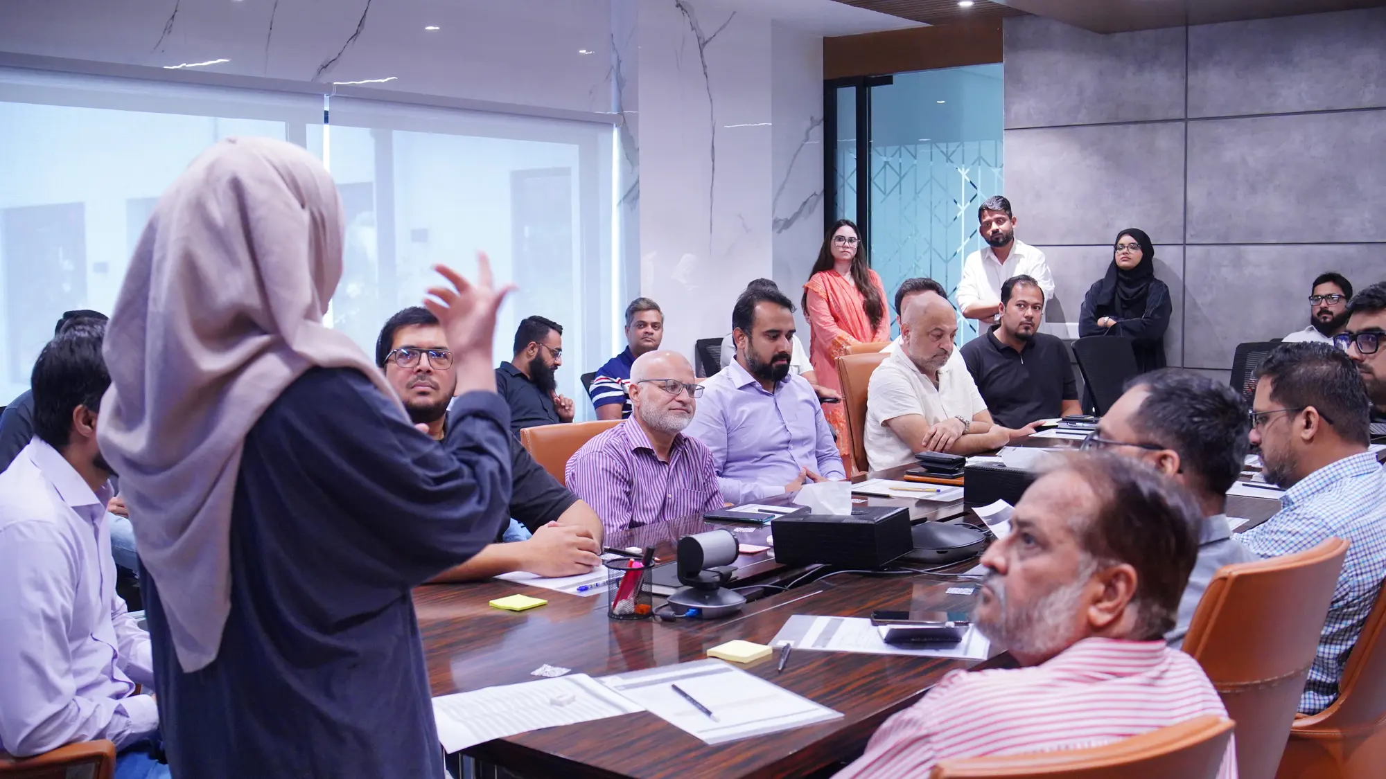 A facilitator in a beige headscarf gesturing as she speaks to Amreli Steels leaders seated around the boardroom table.
