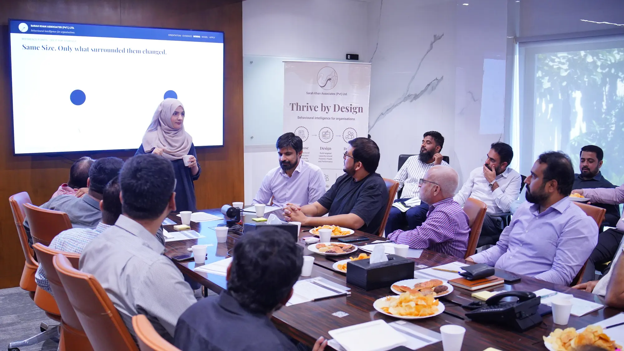 Amreli Steels leaders around a boardroom table in Karachi as a facilitator presents, with a Thrive by Design banner and a session slide behind her.