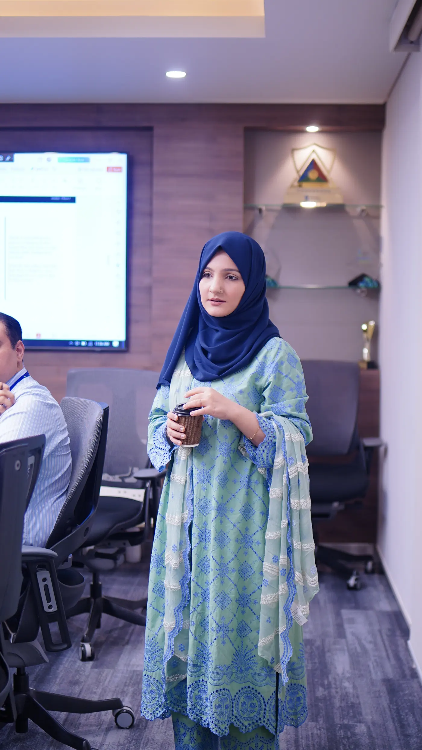 A facilitator in a blue headscarf standing with a cup during the Standard Chartered session, a screen behind her.