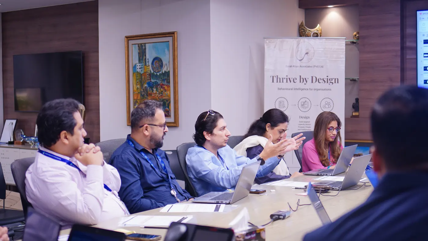 Standard Chartered colleagues seated along a conference table, one woman gesturing as she speaks, with a Thrive by Design banner behind them.