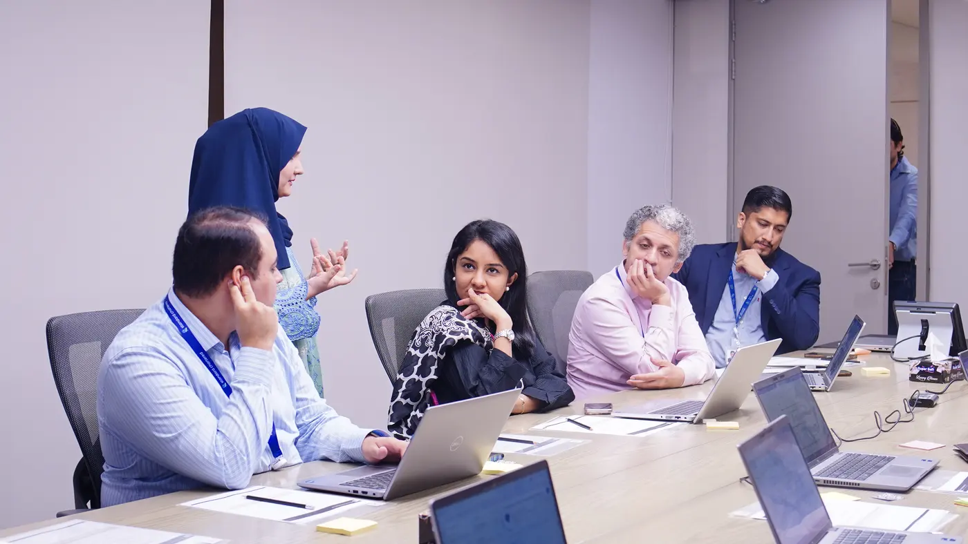 A facilitator in a navy headscarf standing and speaking to Standard Chartered colleagues seated along the conference table.